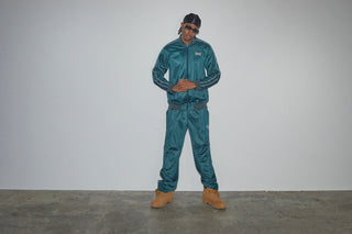 En mand poserer mod en enkel grå væg iført et glansfuldt, mørkt turkis Everlast tracksuit med hvide detaljer langs ærmer og ben. Han kombinerer outfittet med brune støvler, solbriller og en sort durag, hvilket giver et cool og selvsikkert streetwear-look.